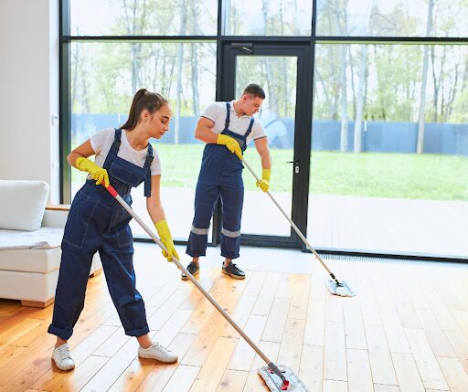 Two people in cleaning uniforms pushing mops over a hardwood floor.